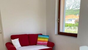 Un sofá rojo con un cojín a rayas coloridos, una alfombra de cuadros y una silla en una habitación con una ventana y una puerta.