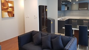 Ein modernes Wohnzimmer mit einem dunklen Sofa und einer Küchenzeile, die einen Kühlschrank, eine Spüle und Esstische mit Stühlen umfasst.