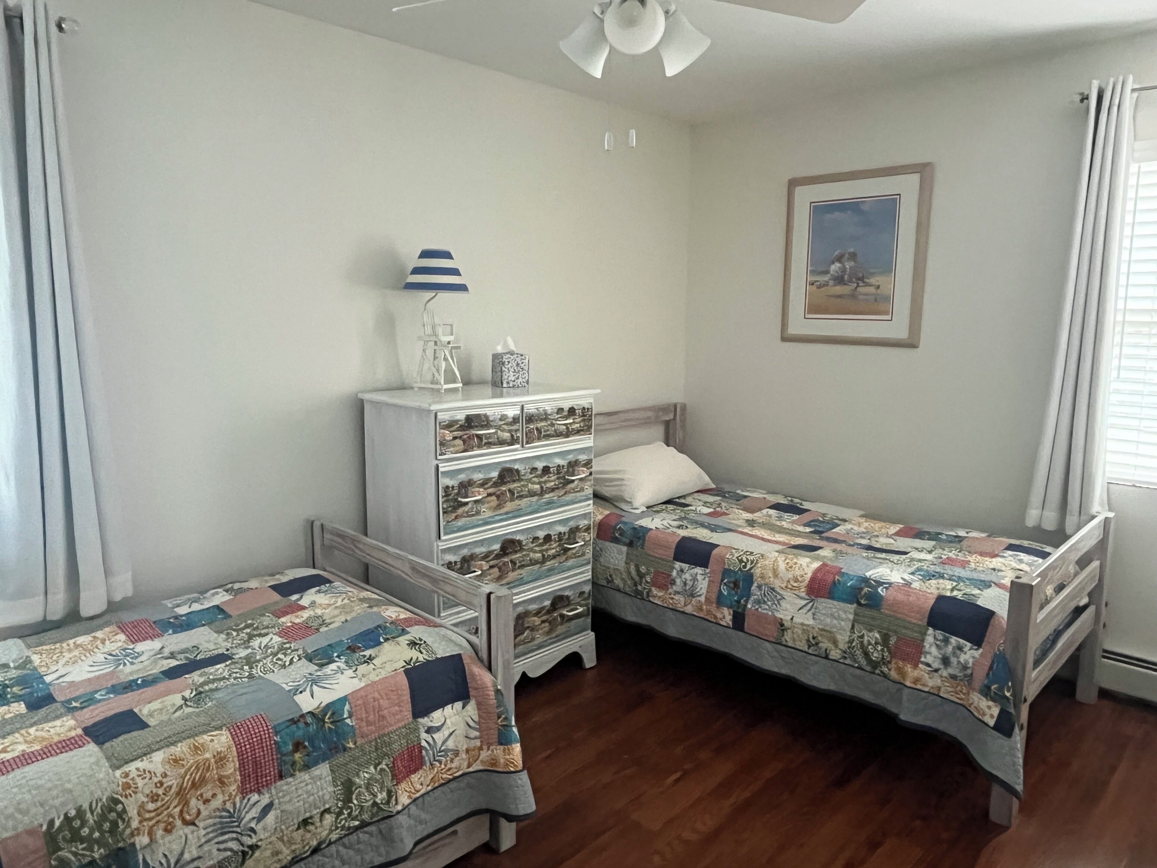 Une chambre avec deux lits simples, une commode avec des tiroirs, un ventilateur de plafond et un tableau encadré au mur.