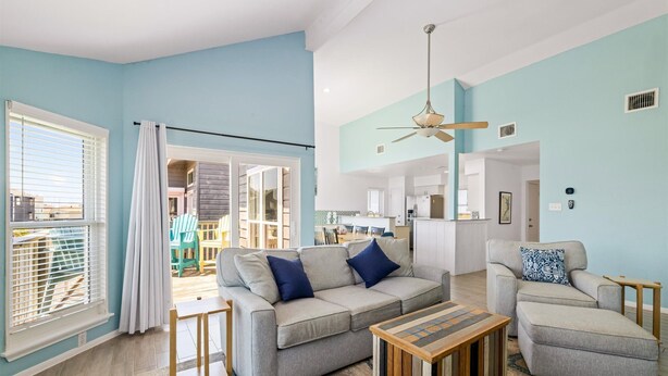 A spacious living room with a grey sofa, a wooden coffee table, and a grey armchair. There is a ceiling fan, a window with blinds, and a view into the kitchen area.