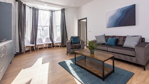 Una sala de estar moderna con un sofá, una mesa de centro y sillas. Hay una zona de cocina con armarios y un refrigerador.