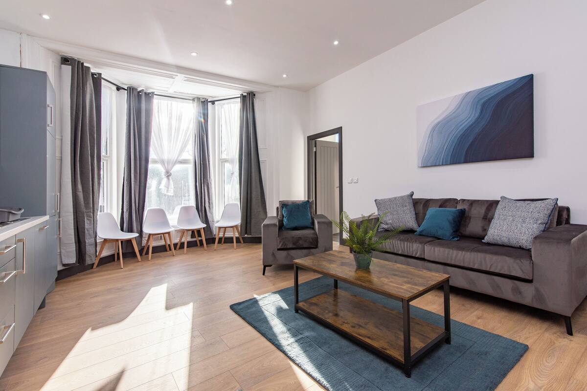 Un salon moderne avec un canapé, une table basse et des fauteuils. On y trouve également un coin cuisine équipé de placards et d’un réfrigérateur.