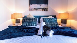 Una cama bien hecha con una colcha azul, almohadas y un par de zapatos sobre la cama. Hay dos lámparas de noche y un cuadro en la pared.