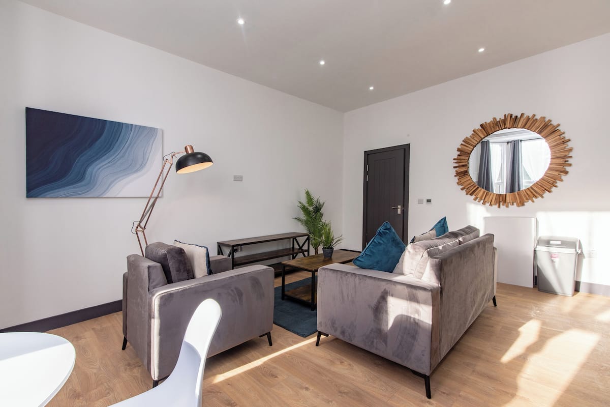 Un salon moderne avec un canapé gris, un fauteuil blanc, une table basse, un lampadaire, un grand tableau abstrait, un miroir et une plante en pot.