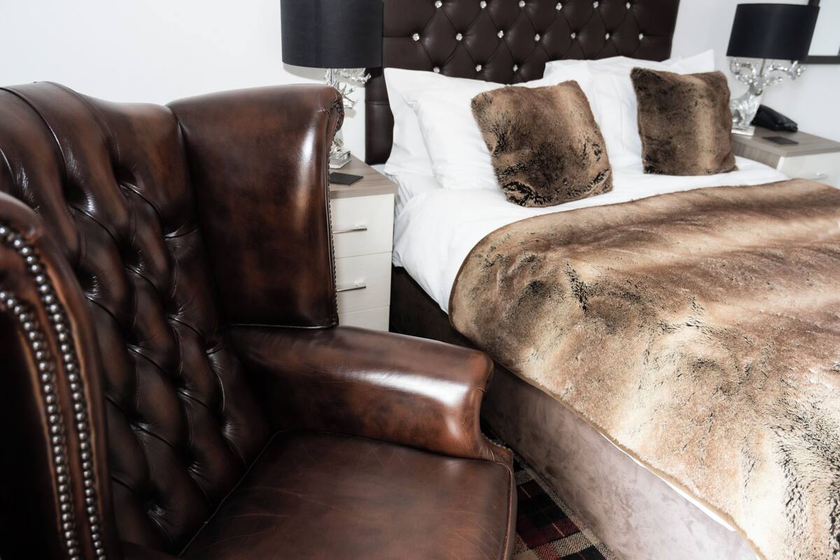 A bedroom with a tufted leather armchair, a bed with a fur throw, and a nightstand with a lamp.