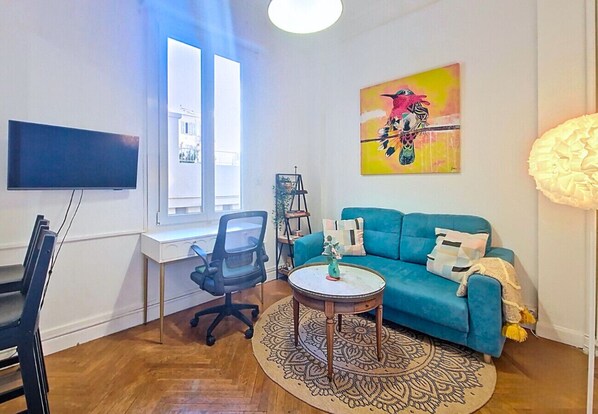A modern living room with a blue sofa, a round coffee table, a desk with a chair, and a large window. - N&J - CLEMENCEAU (Niza)