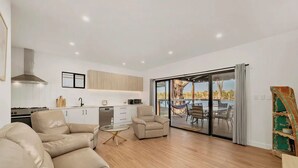 A modern living room with a sofa, armchair, and coffee table. There is a kitchen area with cabinets and a stove. A sliding glass door leads to an outdoor patio with a hammock and dining table. - Black Shack - 13 Paisley Island (Paisley)