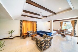 A spacious living area with a sofa, armchairs, a coffee table, and a dining table with chairs. There is a ceiling fan, a potted plant, and a view through large windows. - Country Inn Tarika Riverside Resort Jim Corbett (Ramnagar)