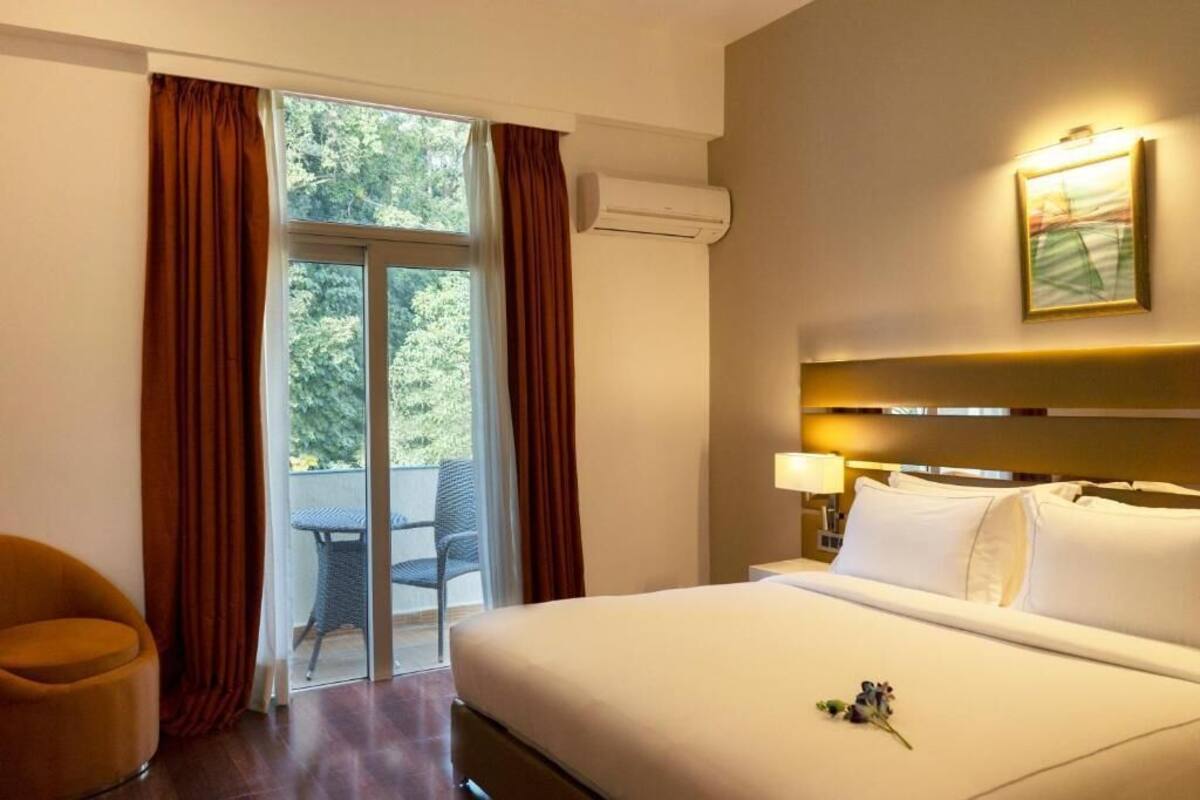 A hotel room with a bed, a chair, a window with curtains, an air conditioning unit, and a wall-mounted light fixture.