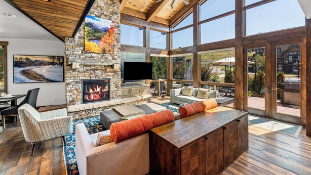 A spacious living room with a stone fireplace, wooden ceiling, and large windows offering a view of the outdoors.