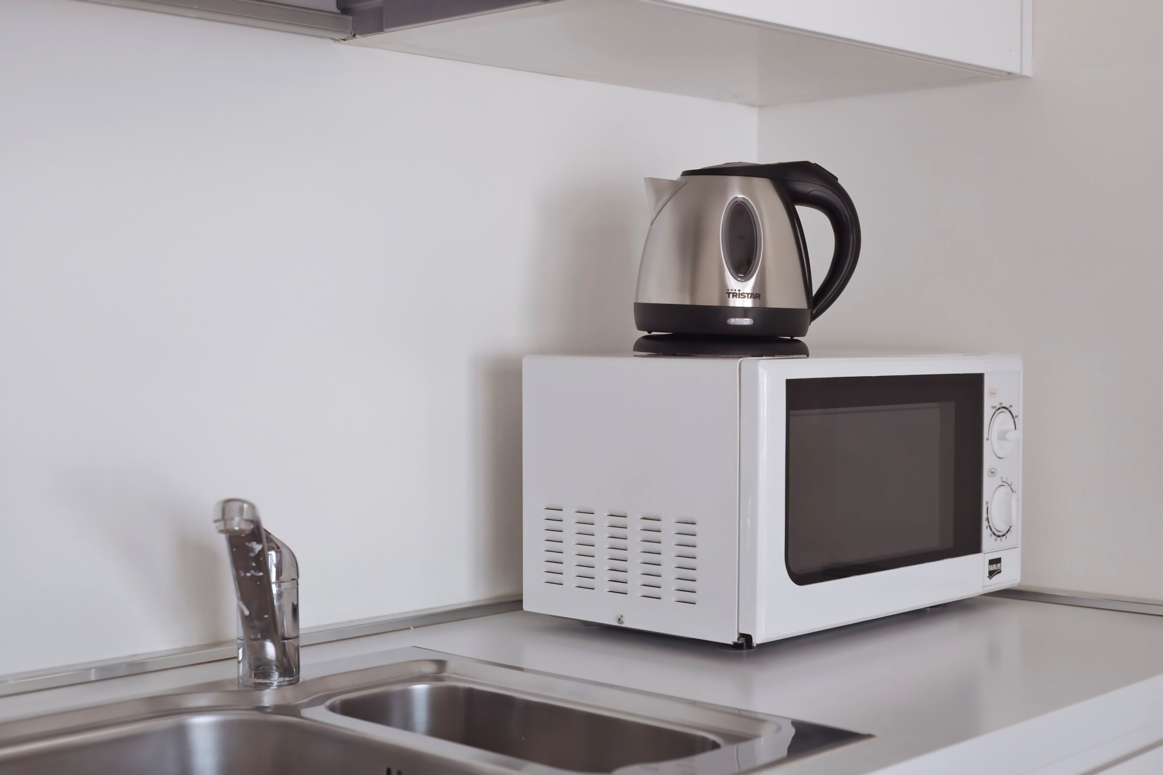 A kitchen with a white microwave, a kettle, and a stainless steel sink.