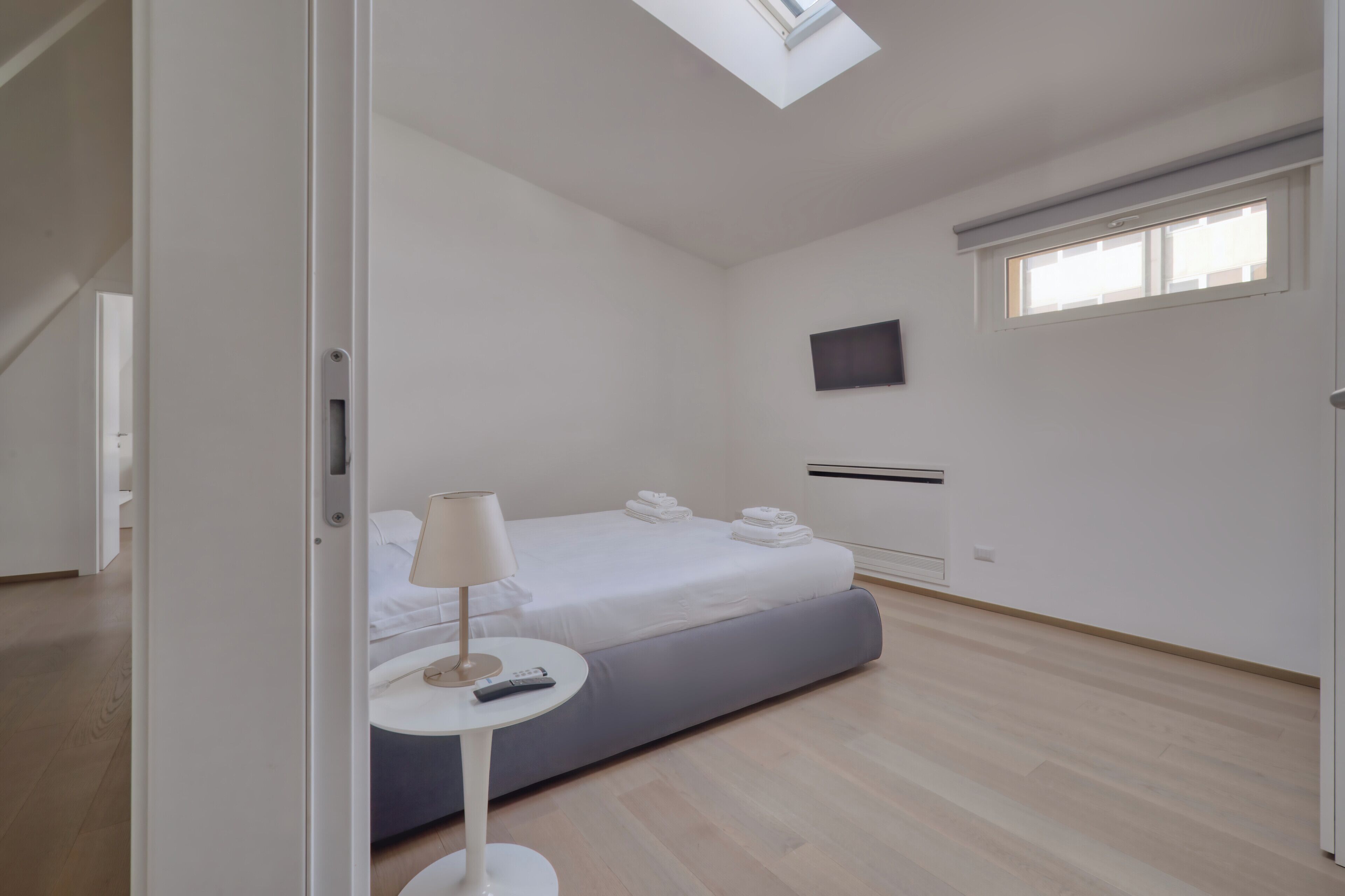 Ein Hotelzimmer mit Bett, Nachttisch mit Lampe, Fernbedienung, Fenster und einem an der Wand befestigten Flachbildfernseher.