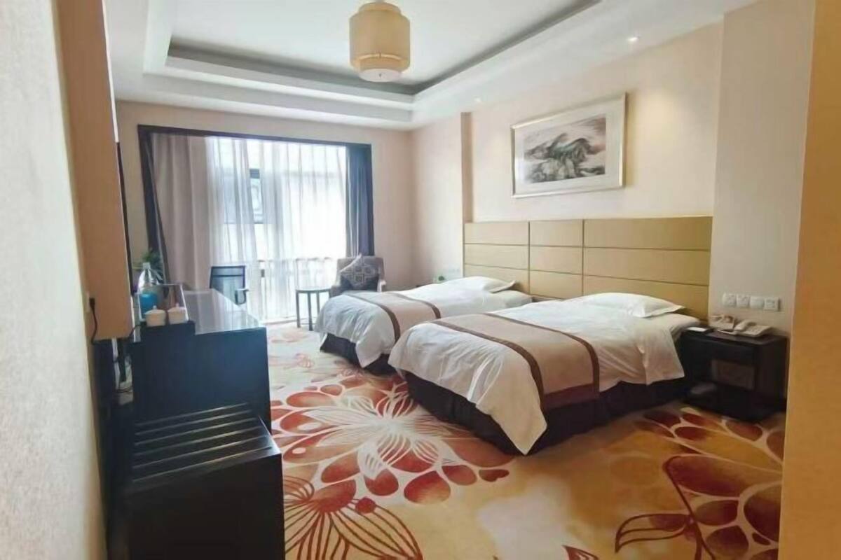 A hotel room with two beds, a large window with curtains, a painting on the wall, and a carpet with a floral pattern.