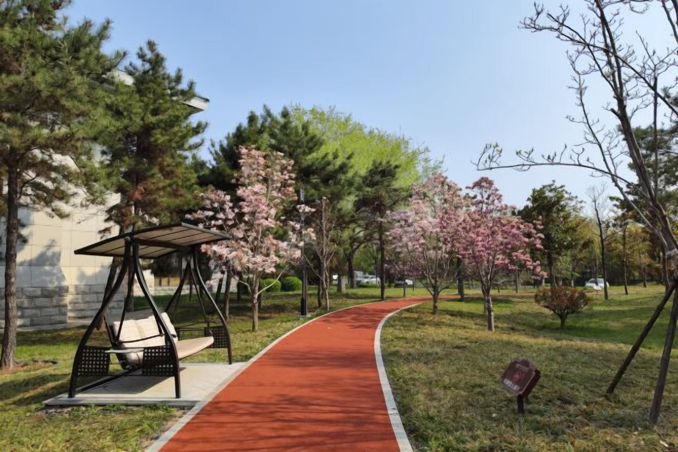 A park pathway with a swing set and a bench, surrounded by trees and a red path.
