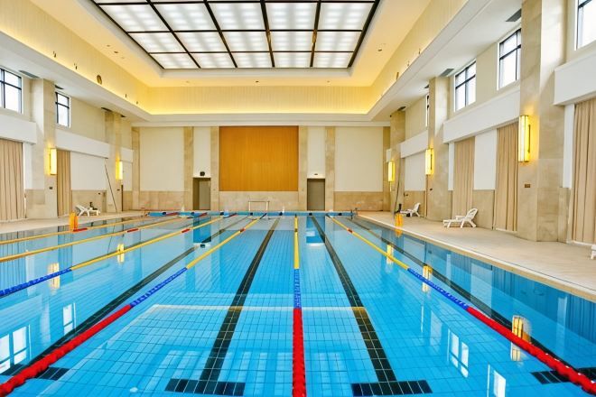 An indoor swimming pool with multiple lanes, a high ceiling, and a seating area along the side.