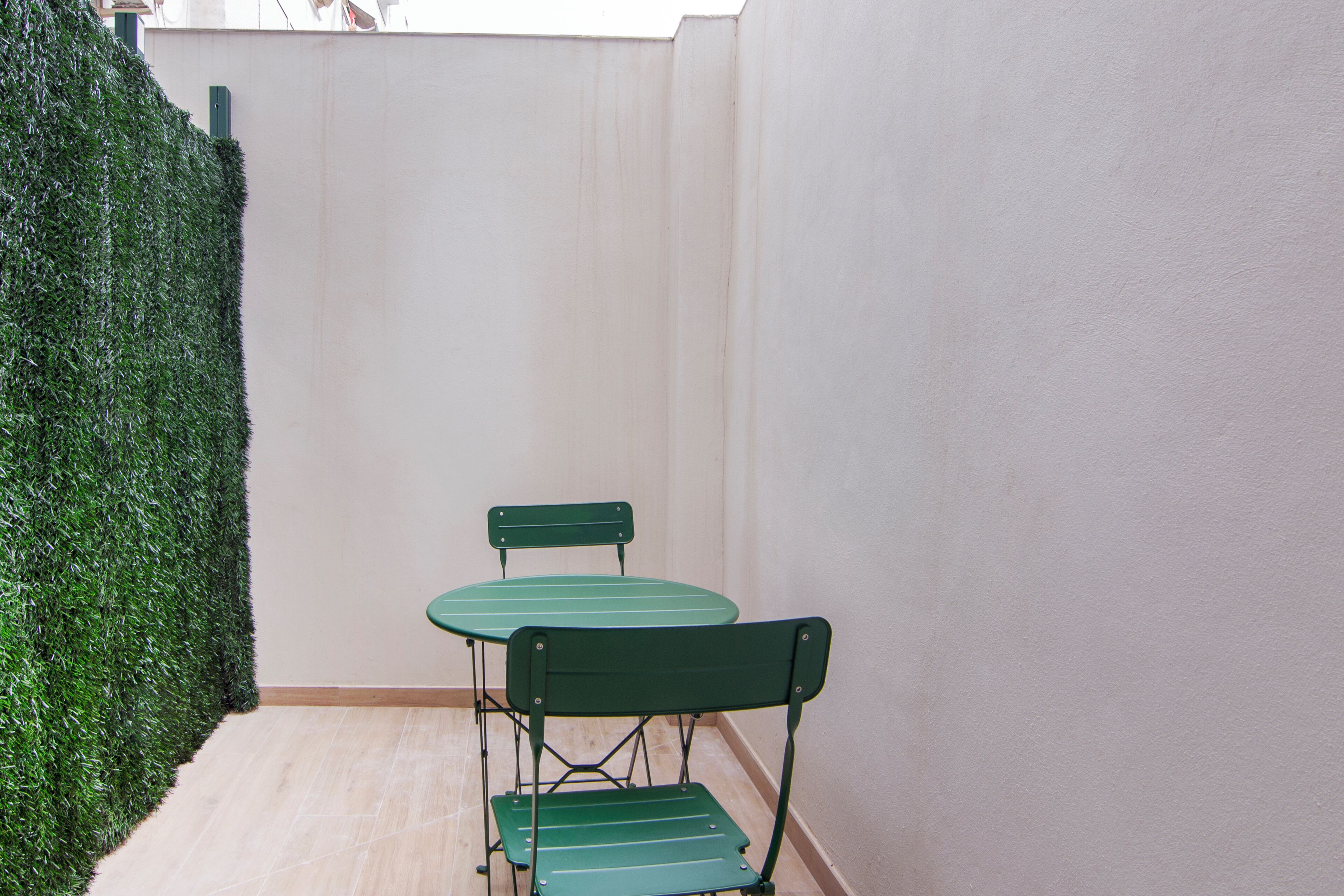 Un pequeño patio amurallado con una mesa verde y dos sillas, rodeado por un muro blanco y un seto verde.