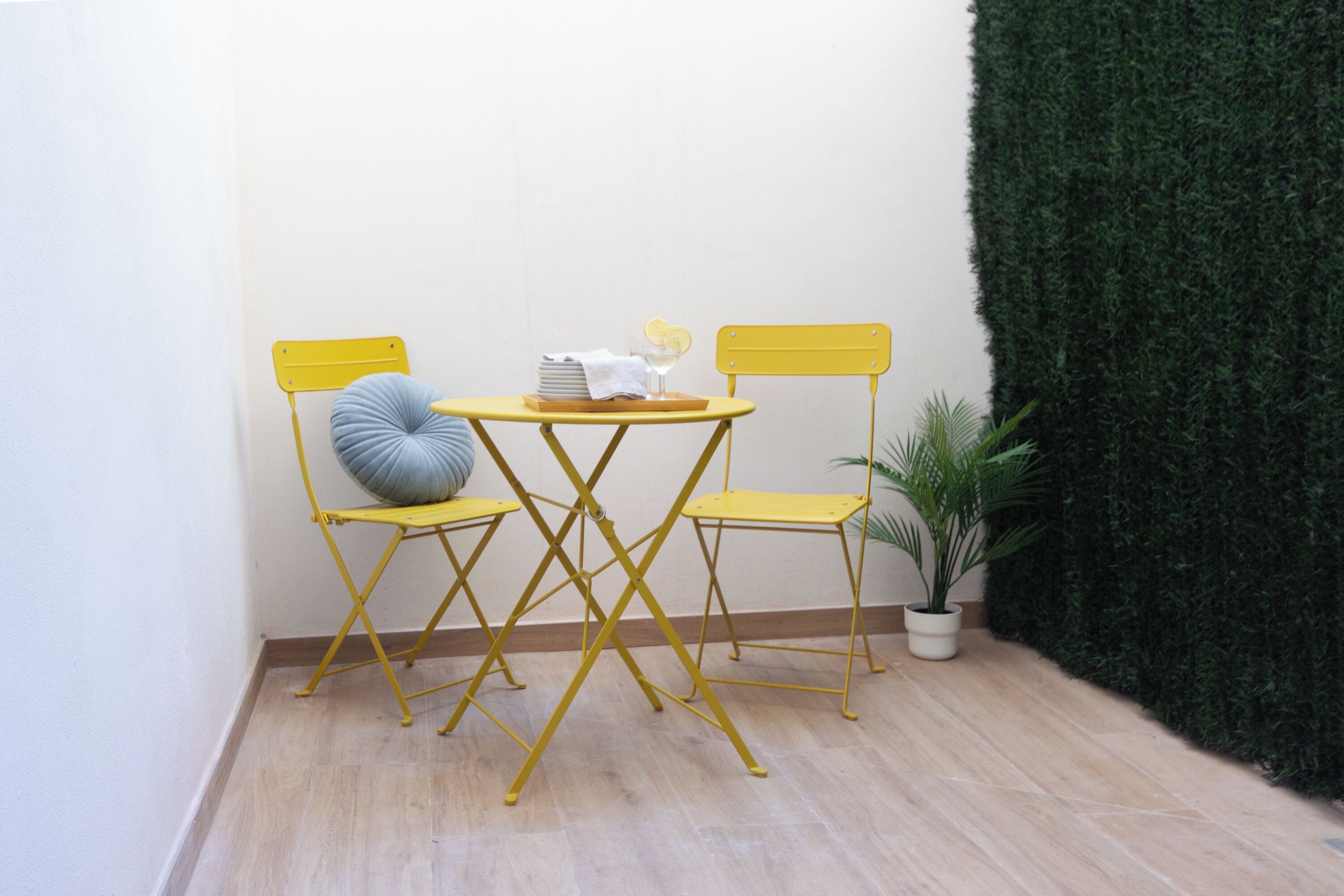 A small, yellow foldable table with two matching chairs, a potted plant, and a neatly arranged table setting including a glass and a striped napkin.