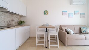 Een moderne keuken met witte kastjes, een eettafel met stoelen, een beige bank en een wandairconditioner.