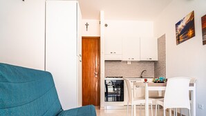 Ein kompakter Wohnbereich mit einem türkisblauen Sofa, einer Essecke mit weißen Stühlen, einer Küche mit weißen Schränken und einer Holztür.