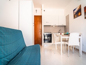 Ein kompakter Wohnbereich mit einem türkisblauen Sofa, einer Essecke mit weißen Stühlen, einer Küche mit weißen Schränken und einer Holztür.