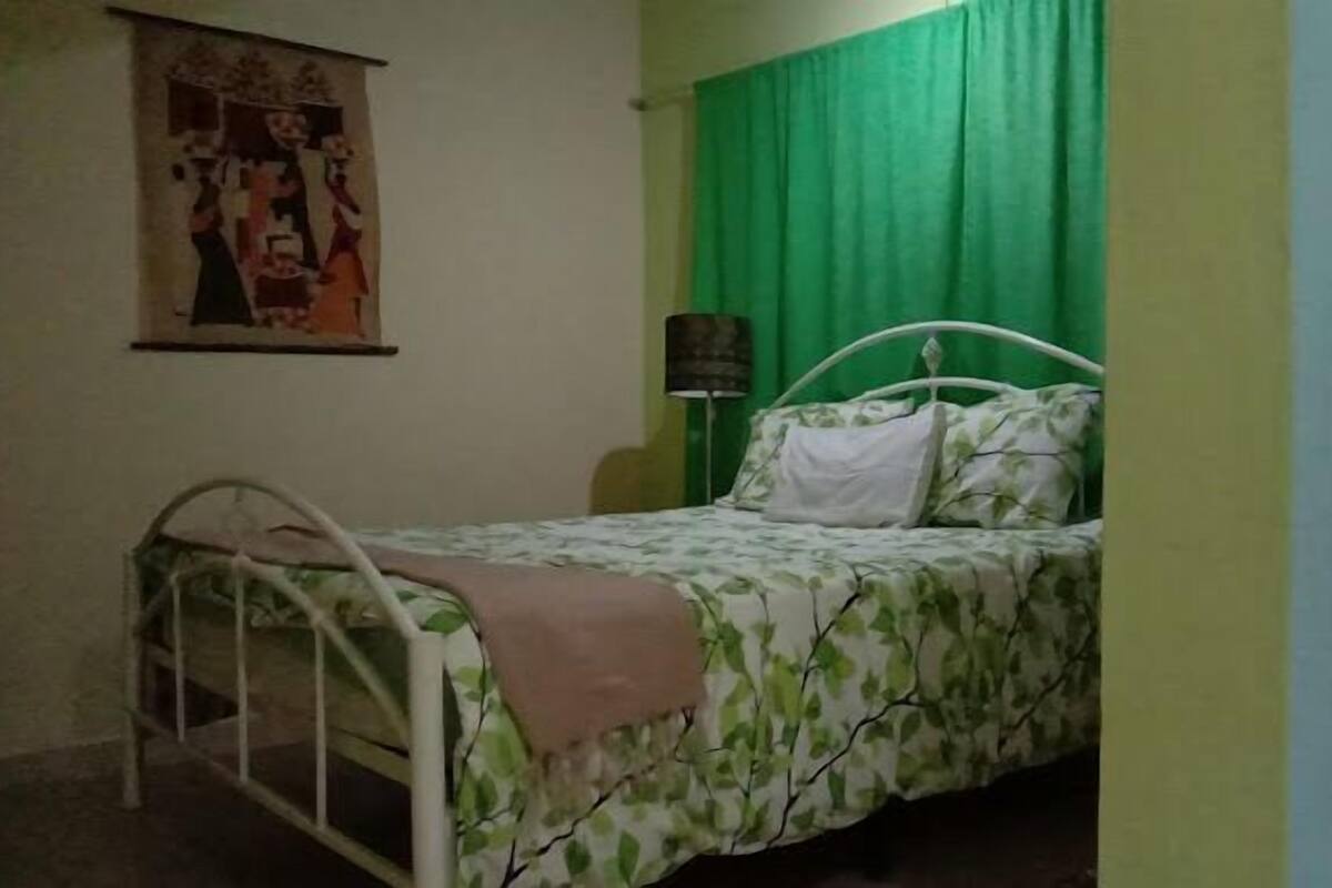 Une petite chambre avec un lit simple, dotée d’un rideau vert, d’un cadre de lit en métal blanc et d’une lampe fixée au mur.