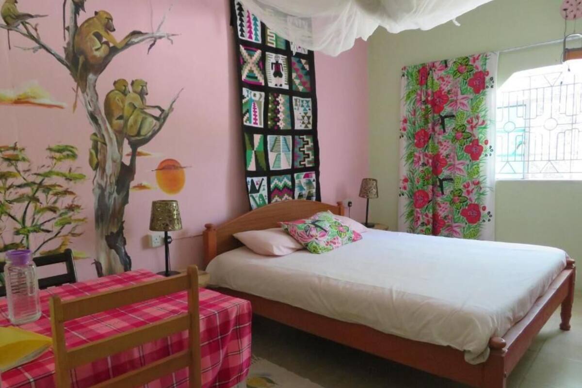 Une chambre à coucher comprenant un lit, une chaise, une table, un mur orné d’une fresque et une fenêtre avec des rideaux.