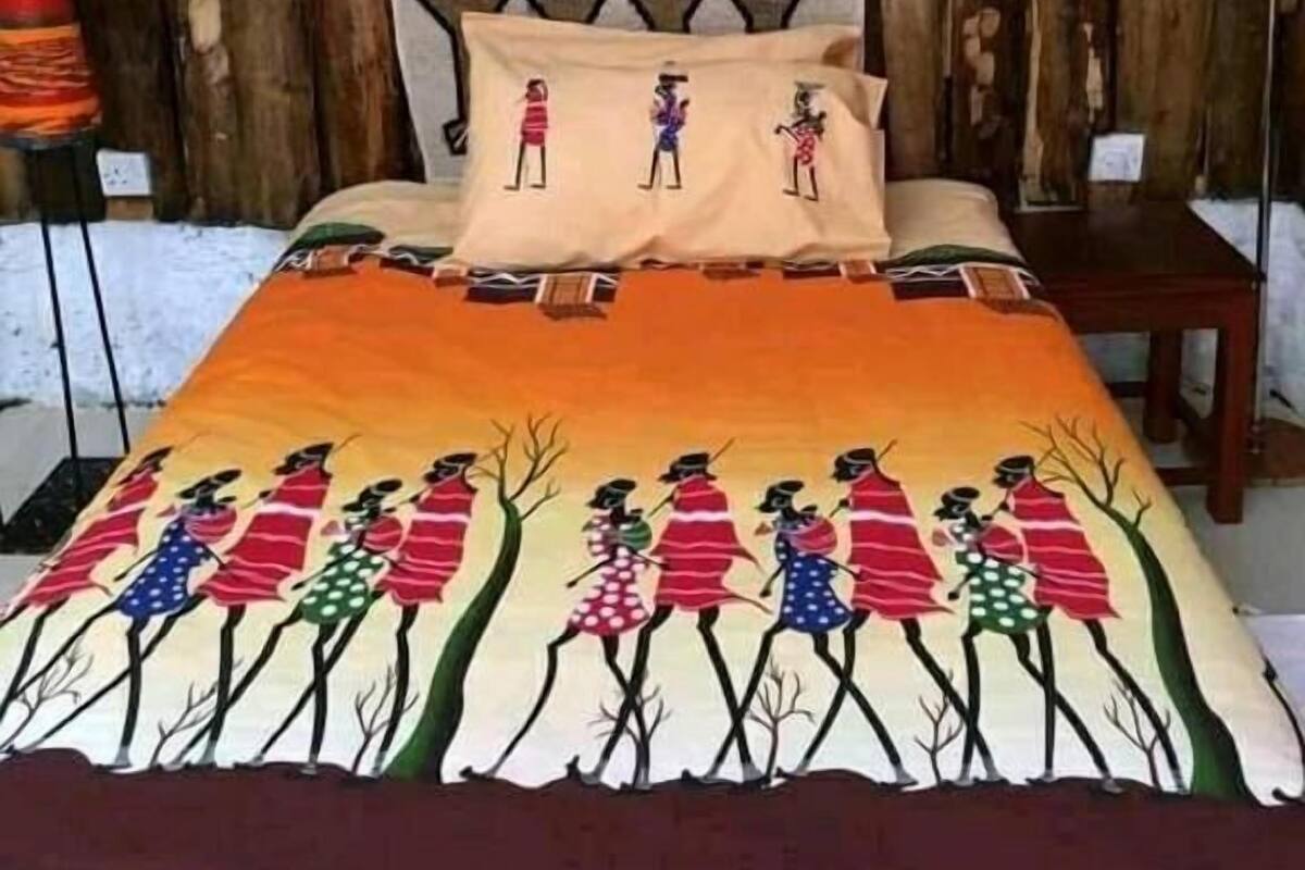 Un lit recouvert d’une couverture colorée ornée de motifs représentant des personnages vêtus de costumes traditionnels, un mur en bois décoré d’œuvres d’art abstraites et une lampe rouge placée à côté du lit.