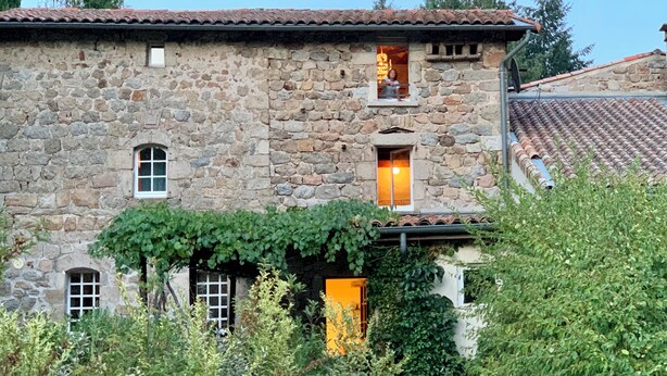A stone building with a tiled roof, a window with a warm light, and greenery around it.