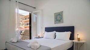 A neatly made bed with white linens and a blue headboard, a small bedside table with a lamp, and a view of a stone building through an open door. - Captain Antonis - Authentic Seafront Living in Gerolimenas (Gerolimenas)