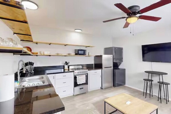 A compact kitchen with a built-in oven, a microwave, and a refrigerator. There is a ceiling fan, a TV mounted on the wall, and a small dining table with two stools.