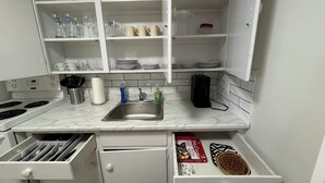 A compact kitchen with white cabinets, a stainless steel sink, and a microwave. There are shelves above the sink holding various kitchen items, and a drawer below the sink containing utensils and a box of cookware. - Cozy 2-Bed Apartment in Town (Wolfville)