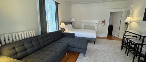 Une chambre d’hôtel moderne avec un canapé d’angle, un lit simple, une petite table avec des chaises et une tête de lit blanche.
