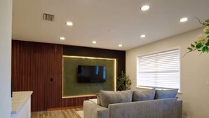 A modern living room with a grey sofa, wooden flooring, recessed ceiling lights, and a flat-screen TV mounted on a wall with a wooden panel design.