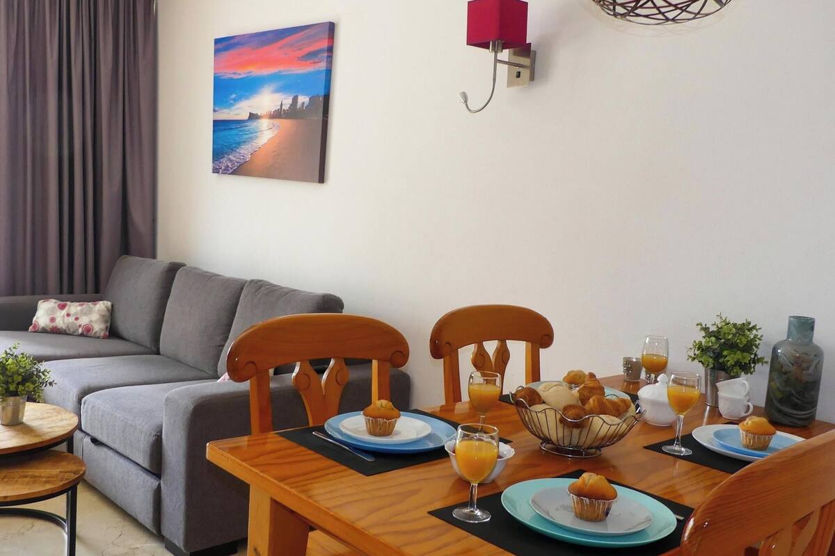 Ein Essbereich mit Holztisch, gedecktem Frühstücksservice, einer grauen Couch und einem Gemälde, das einen Sonnenuntergang über dem Meer zeigt.