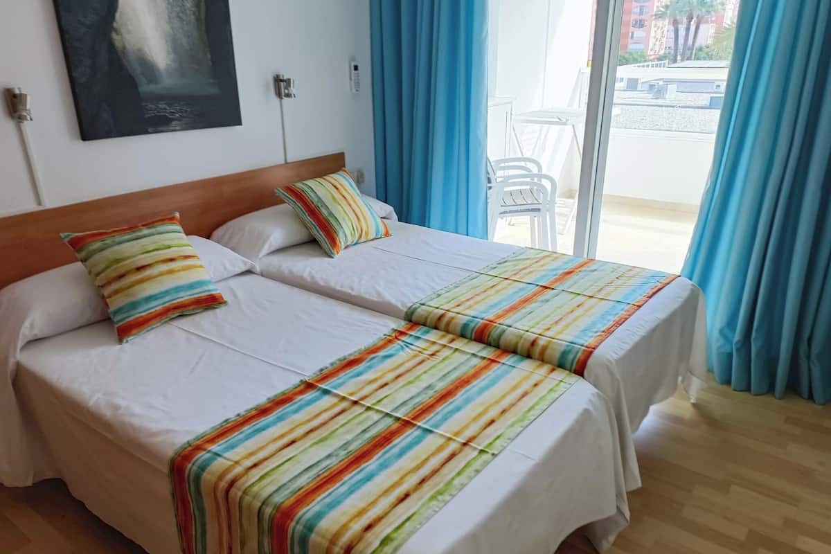 Ein Hotelzimmer mit zwei Betten, einem hölzernen Kopfteil, weißer Bettwäsche, gestreifter Decken und Blick durch eine Glastür nach draußen.