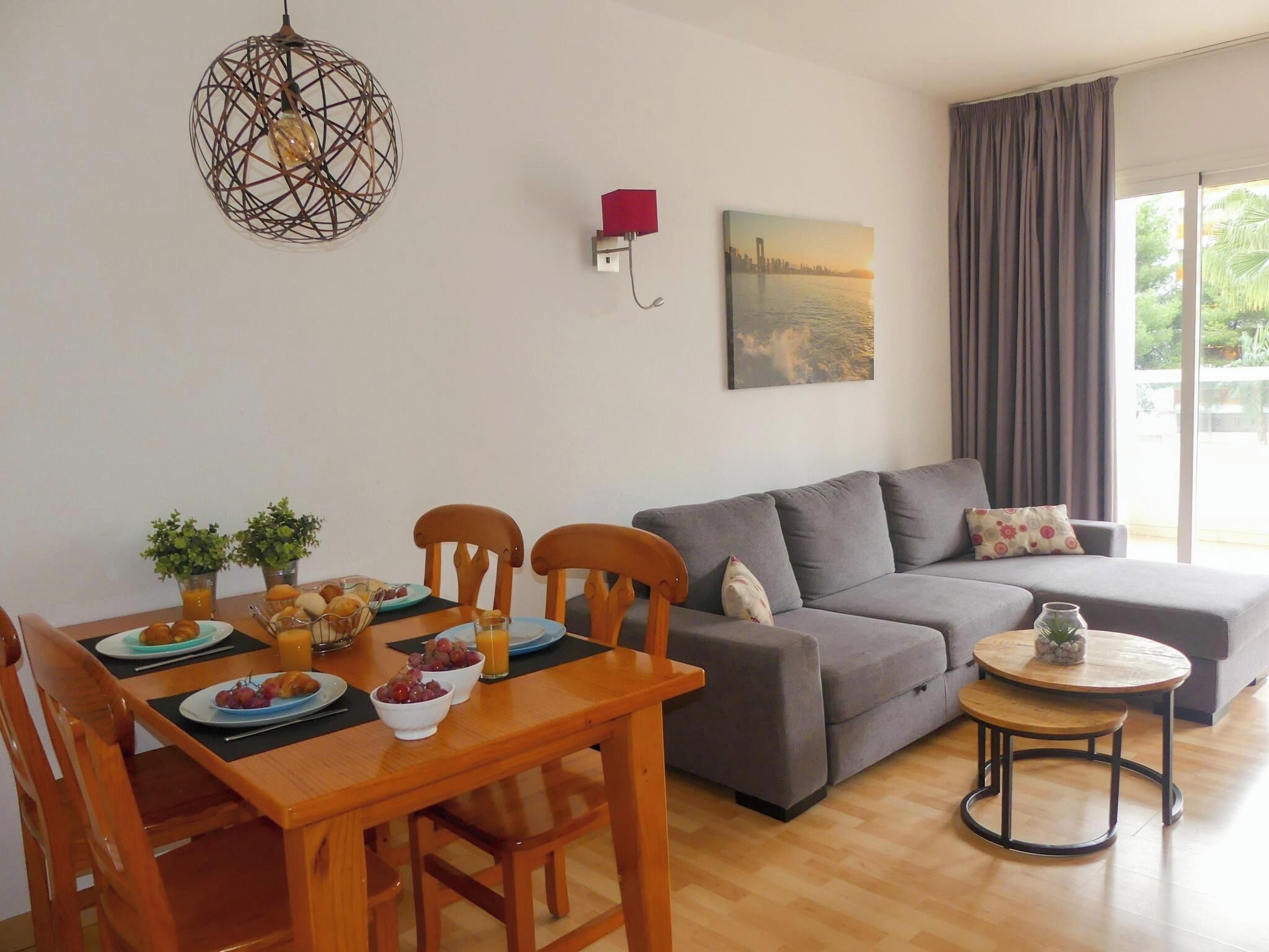 A modern living room with a dining table, wooden chairs, a grey sofa, a small round table, and a hanging light fixture.