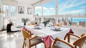 A dining area with a table set for two, large windows with a cityscape view, and a modern chair design. - Siroco-Gemelos 22 (Benidorm)