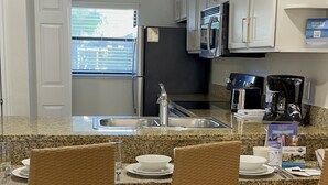 A kitchen with a granite countertop, white cabinets, a black refrigerator, and a coffee maker. There are wicker chairs with cushions. - Cozy 2-bedroom condo with WiFi, AC in Orlando - Close to Universal and Disney (Orlando)