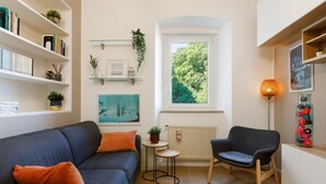 Un saló acollidor amb un sofà blau, una taula rodona petita, una cadira, prestatges amb llibres i plantes, una finestra amb vistes a arbres i un radiador.