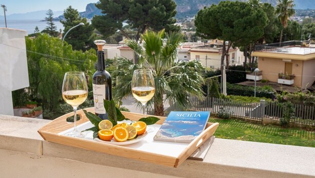 A tray with a bottle of wine, two glasses of wine, and a book titled "Sicilia" on a balcony overlooking a coastal area with houses and trees.