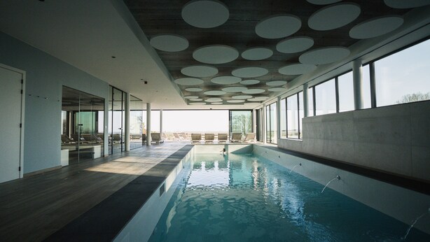 Une piscine intérieure moderne, dotée de grandes baies vitrées offrant une vue dégagée sur le ciel, d’un plancher en bois et d’un plafond orné de motifs circulaires.