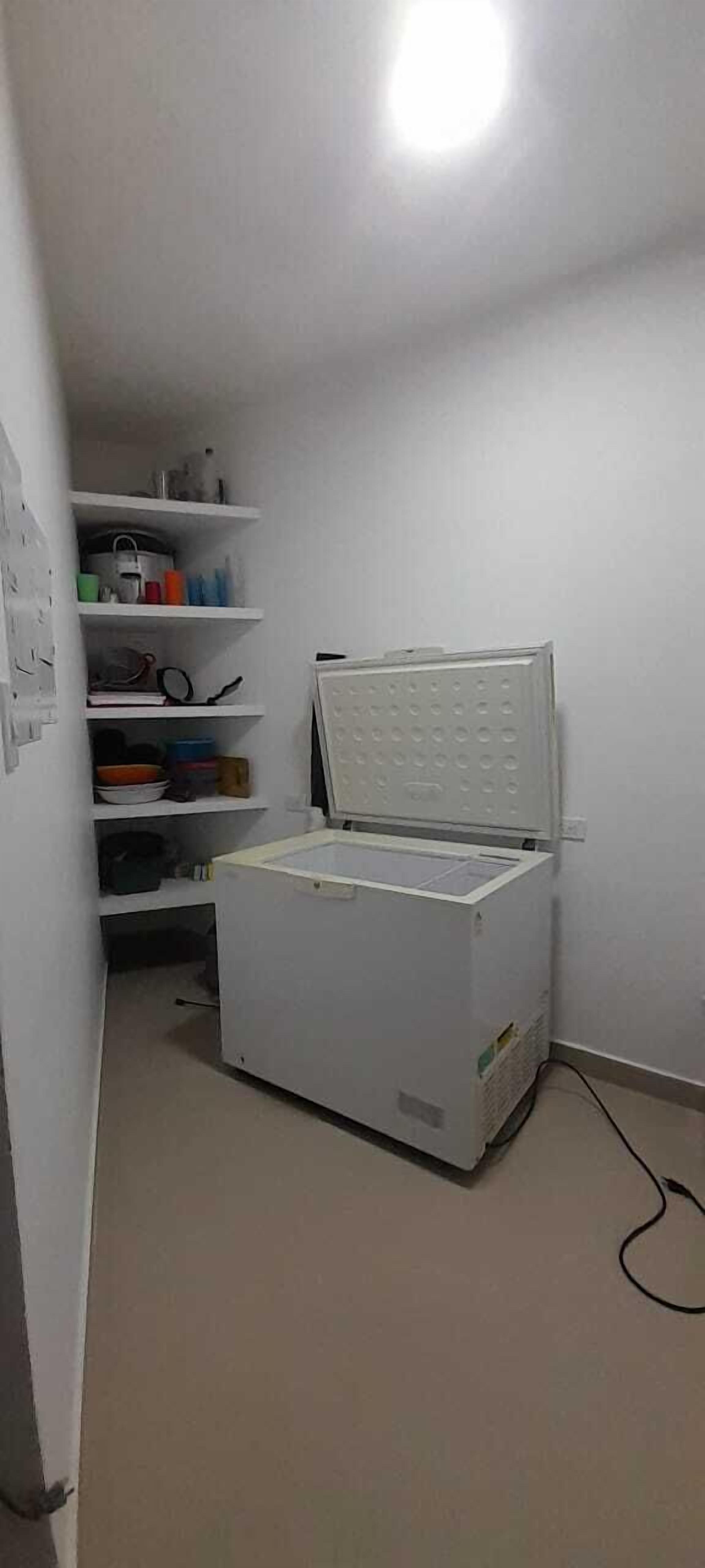Una stanza piccola con un frigorifero bianco, uno scaffale con diversi oggetti e una presa a muro.