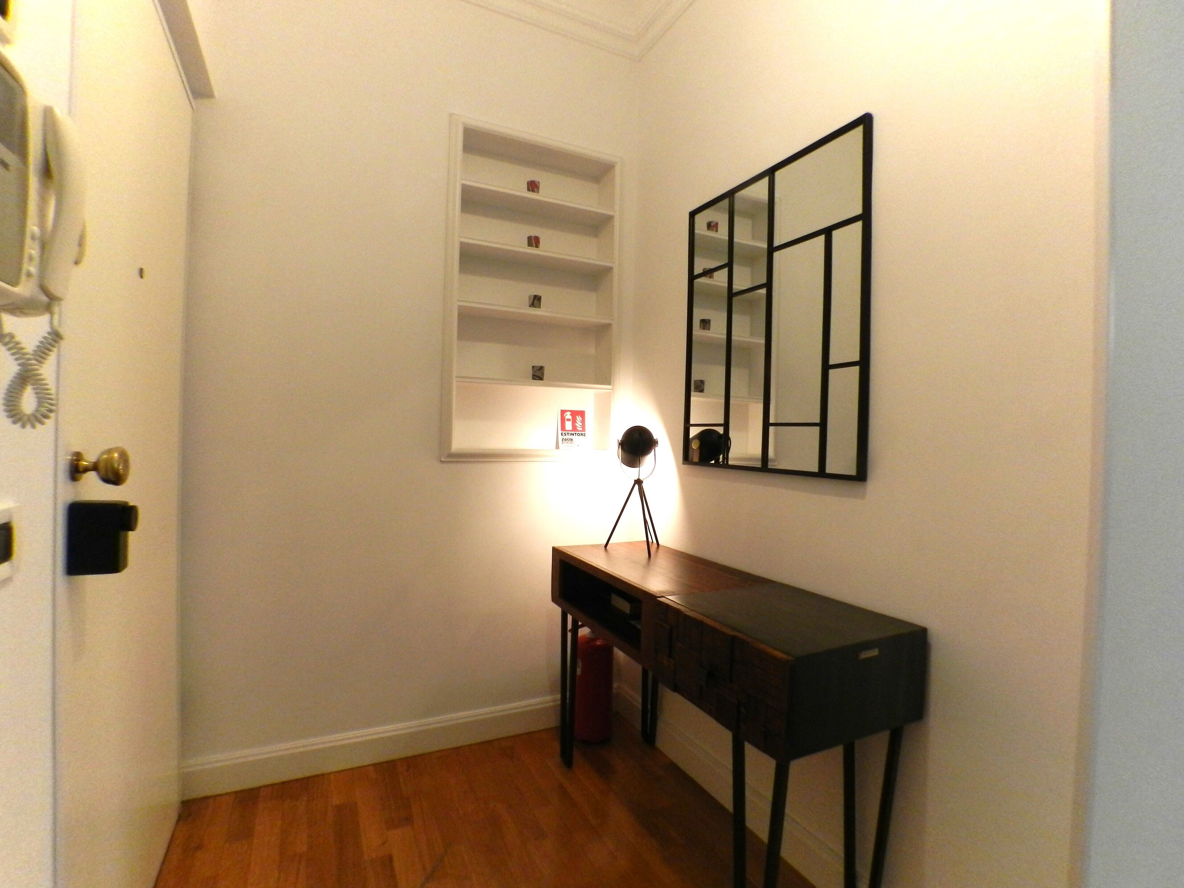 A hallway with a wooden floor, a desk with a lamp, a shelf unit, and a mirror on the wall.