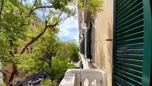 Una calle estrecha con un edificio a la derecha, que presenta un persiano verde y un balcón con barandilla blanca.