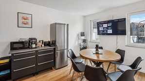A modern kitchen with a stainless steel refrigerator, a microwave, and a coffee machine. There is a dining table with chairs and a TV mounted on the wall displaying a streaming service interface. - Welcome to our stylish new construction vacation home in Dasing! (Dasing)