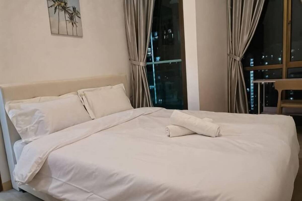 A neatly made bed with white linens, a window with curtains, and a cityscape view at night.