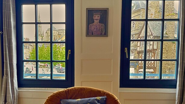 Una habitación con un sillón marrón, una obra enmarcada sobre un ventanal y vista a la calle a través del ventanal.