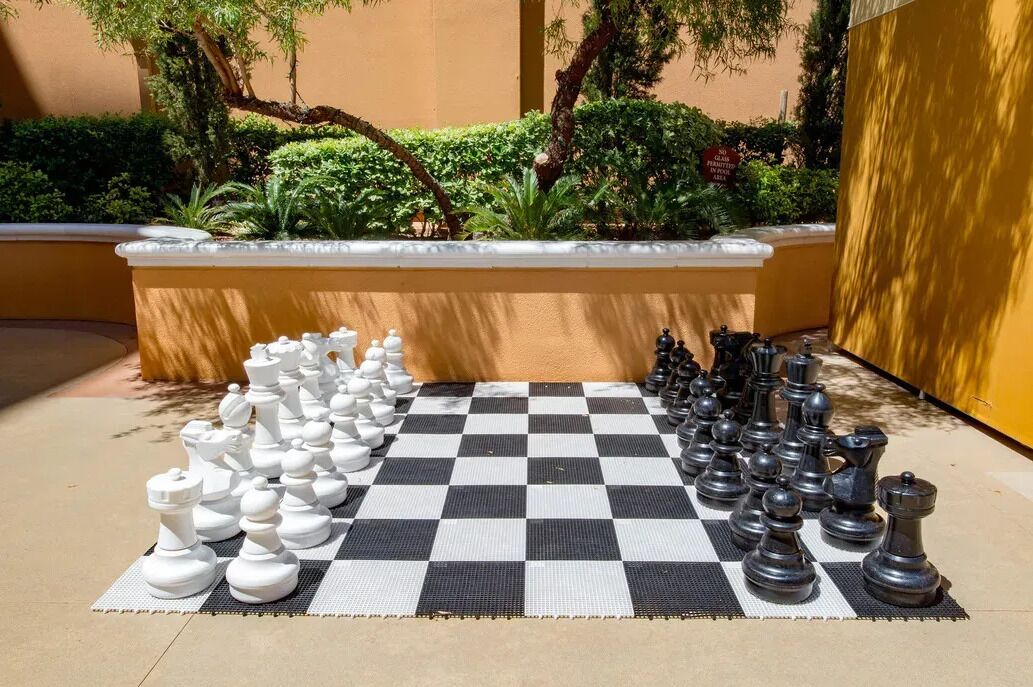 Un grand échiquier installé à l’extérieur, dans une cour dont le sol est à carreaux, avec les pièces d’échecs disposées pour une partie.