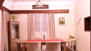 Un comedor con una mesa de madera, sillas color beige, un camino de mesa rojo y una ventana con cortinas estampadas.