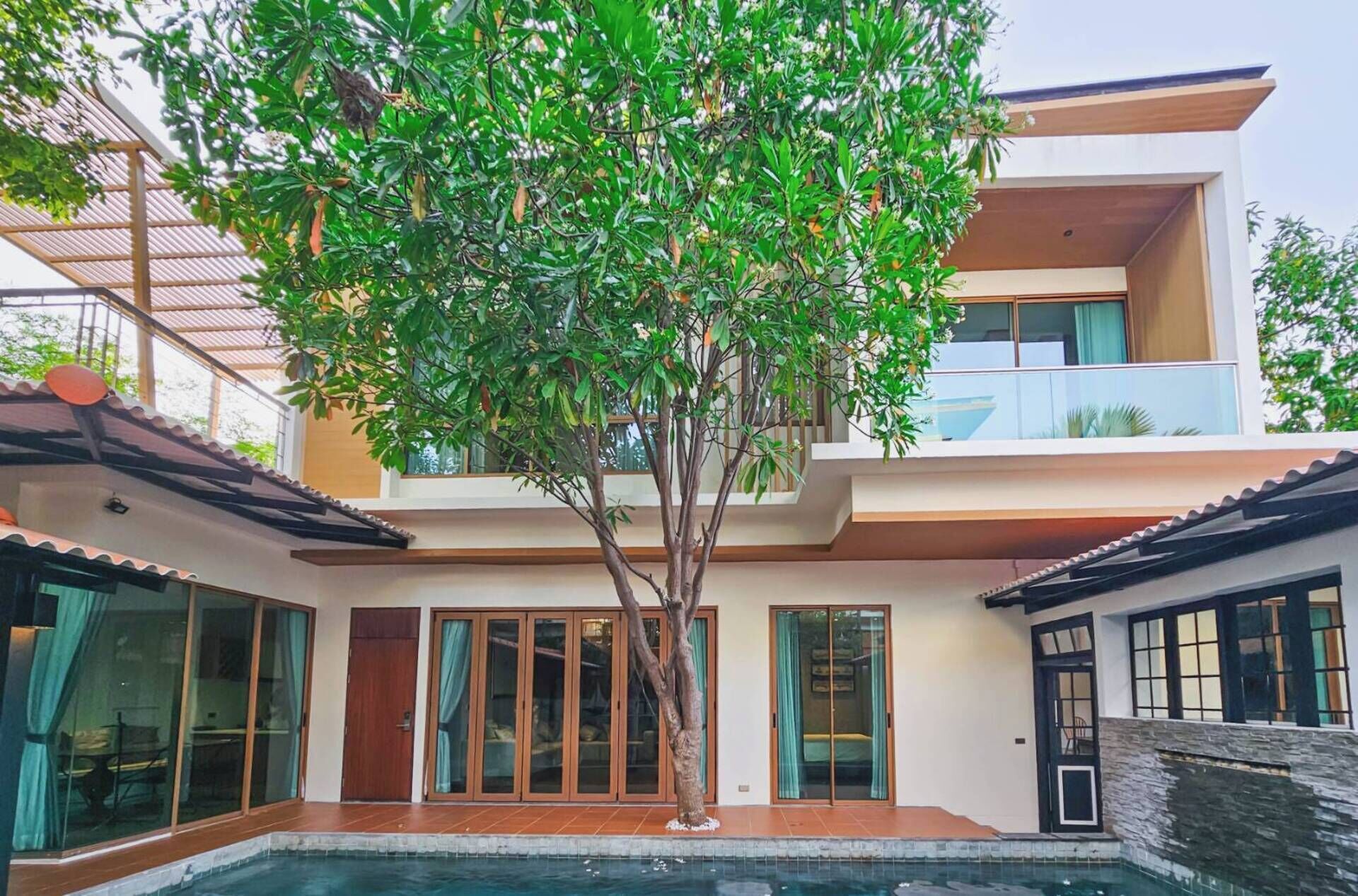 A modern multi-story house with a swimming pool, large windows, and a tree in the courtyard.